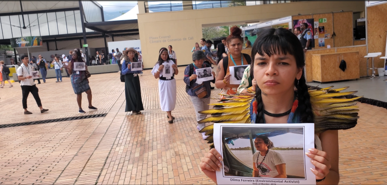 A line of people holding up black-and-white pictures of environtmental defenders who were killed or disappeared. The first person is an indigena, wearing a cape of yellow feathers.